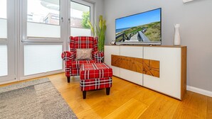 En stue med en rød ternet sofa, et matchende fodskammel, et fjernsyn på et hvidt skab og et stort vindue med persienner.