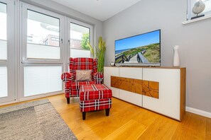 Una sala de estar con un sofá rojo a cuadros, un reposapiés a juego, un televisor sobre un mueble blanco y un ventanal con persianas.