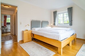 A bedroom with a wooden bed, a bedside table with a lamp, a window with curtains, and a door leading to another room. - Kate Kleinod - Neu!!! Ab September 2022 in der Vermittlung. - Schönes Reihenendhaus in Ruhiger Lage (Sylt-Ost)