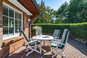 A patio with a round table and four striped chairs, a brick wall, and a window overlooking a hedge.
