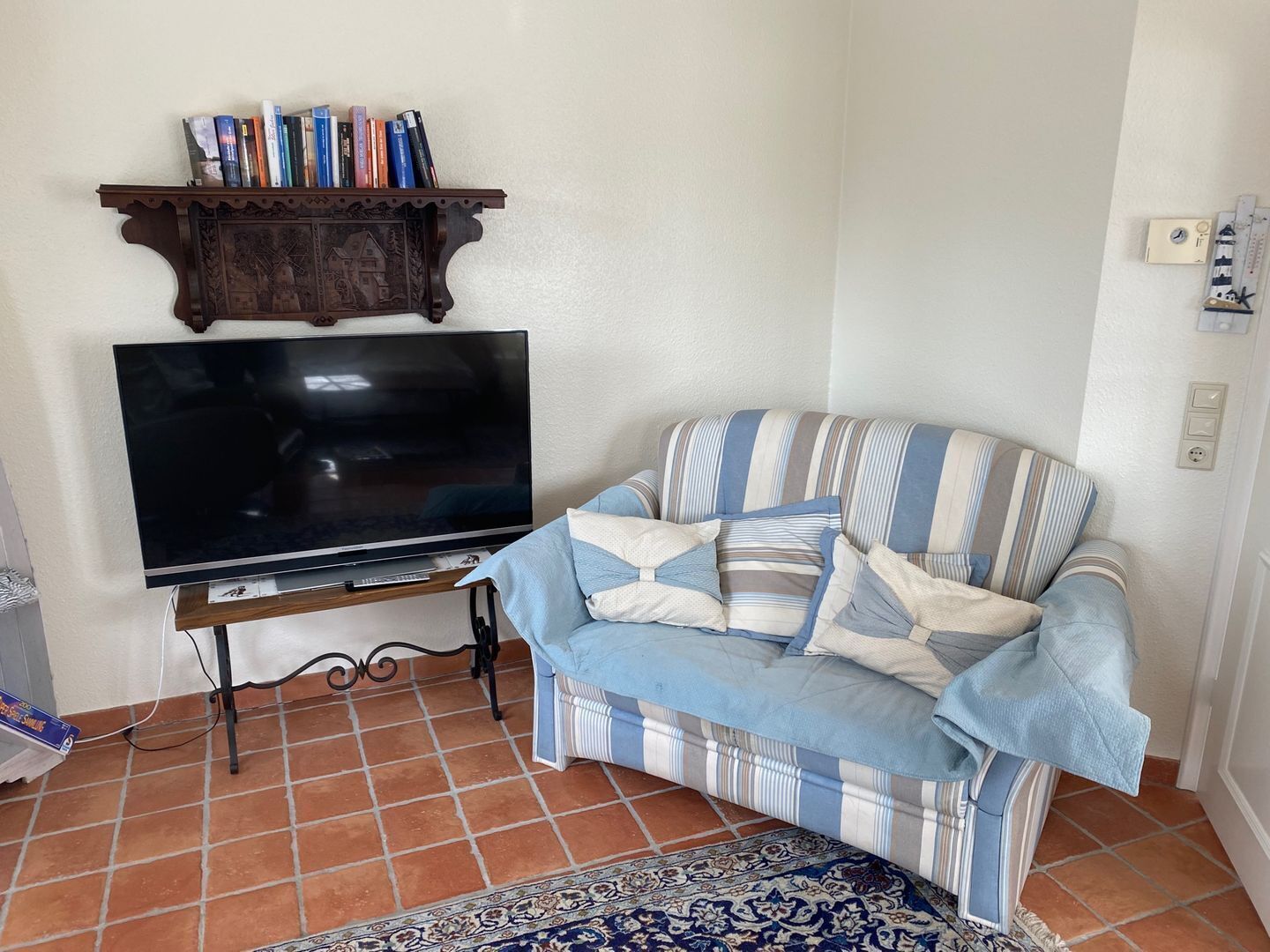 A living room with a flat-screen TV, a striped sofa, a patterned rug, and a wooden shelf with books.