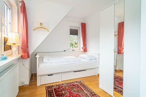 A bedroom with a white bed, a red patterned rug, a window with pink curtains, and a mirror on the wall.