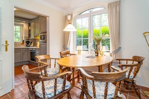 A dining area with a round wooden table and chairs, a pendant light, and a view of the kitchen and garden through large windows. - Kate Kleinod - Neu!!! Ab September 2022 in der Vermittlung. - Schönes Reihenendhaus in Ruhiger Lage (Sylt-Ost)