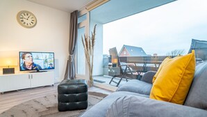 Ein modernes Wohnzimmer mit einem grauen Sofa, einem schwarzen Hocker und einem gelben Kissen. Ein Fernseher steht auf einer weißen Kommode, und es gibt ein Fenster mit Blick auf Häuser.