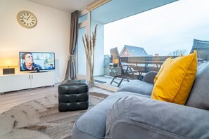 A modern living room with a grey sofa, a black ottoman, and a yellow pillow. A television is on a white cabinet, and there is a window with a view of houses outside.