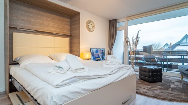 Ein modernes Hotelzimmer mit einem großen Bett, einem Holzkopfstück, einer Wanduhr, einem Fernseher und einem Balkon mit Blick auf Gebäude.