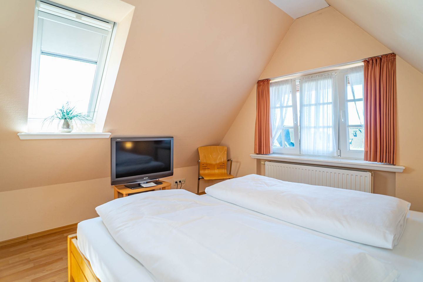 Ein Schlafzimmer mit einem Bett, einem Fernseher auf einem kleinen Tisch, einem Sessel und einem Fenster mit Vorhängen.