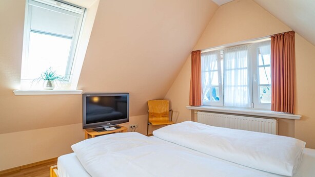Une chambre à coucher avec un lit, un téléviseur posé sur une petite table, une chaise et une fenêtre avec des rideaux.