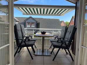 Um alpendre coberto com duas cadeiras, uma mesa e um banquinho de metal, e vista para casas com telhados de cerâmica.