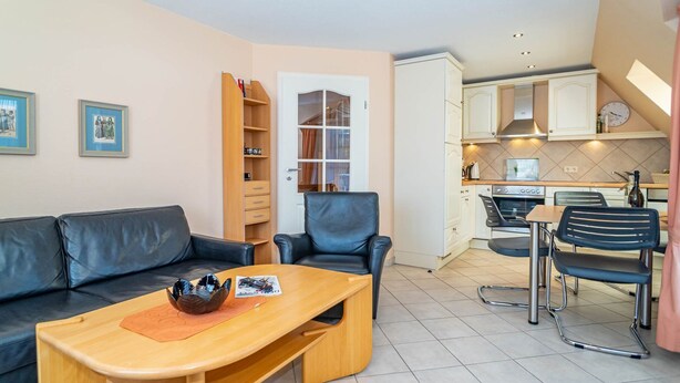 A living room with a sofa, a coffee table, and a dining area with chairs. There is a kitchen in the background with cabinets and a stove. - Ammerland Whg. 2 - Spacious 3-room apartment for up to 4 persons, with balcony. (Sylt)