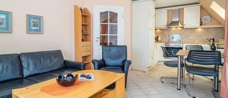 Un salon comprenant un canapé, une table basse et un coin repas avec des chaises. Au fond, on aperçoit une cuisine avec des placards et une cuisinière.