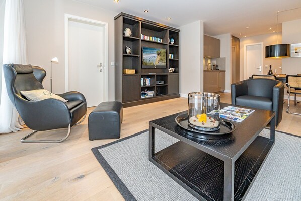 Un salon moderne avec un canapé noir, un pouf noir, une table basse en verre sur laquelle sont posés des magazines, et une bibliothèque contenant divers objets.