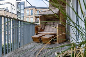 Un balcon doté d’une terrasse en bois, d’un espace salon avec des coussins et d’un auvent, et offrant une vue sur les bâtiments voisins.