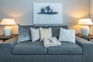 A grey sofa with four cushions, two side tables with lamps, and a painting on the wall.