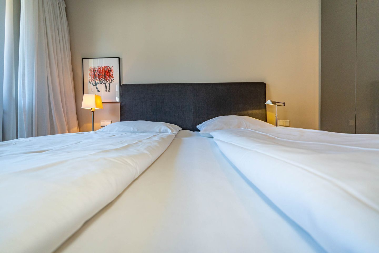Et hotelværelse med to senge, en mørk sengegavl, et billede på væggen og en lampe på hver side af sengen.