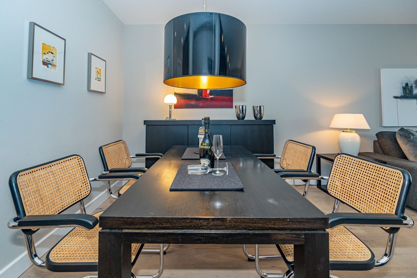 Ein zeitgemäß eingerichteter Essraum mit einem langen Tisch, sechs Stühlen, einem Kronleuchter in der Mitte und einer Couch im Hintergrund.