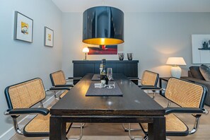 Une salle à manger moderne avec une longue table, six chaises, un lustre central et un canapé en arrière-plan.