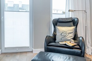 A black leather armchair with a cushion that has "Westbourne House" embroidered on it, positioned next to a window with blinds, a floor lamp, and a book on the armrest.