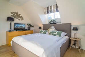 A bedroom with a bed, a wooden dresser, a television, a potted plant, and a window with blinds. - Maarwiet Hüs - Holiday home (detached house) for up to 6 persons with garden and (Wenningstedt-Braderup (Sylt))