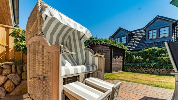 Un espace extérieur abrité avec des sièges, comprenant une chaise en osier et un banc, un mur en pierre et, au fond, une maison en bois.