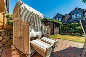 A covered outdoor seating area with a wicker chair and a bench, a stone wall, and a wooden house in the background. - Ammerland Whg. 1 - Spacious vacation apartment for up to 4 persons with garden. (Sylt)
