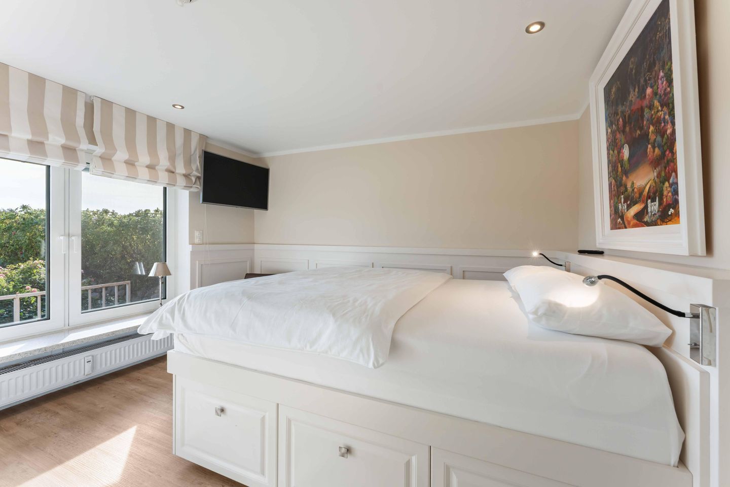 A bedroom with a large bed, a TV mounted on the wall, and a window with a view of trees.