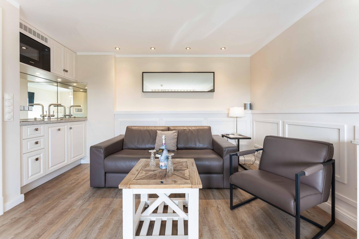 A modern living room with a sofa, armchair, coffee table, and a kitchen area with cabinets and a microwave.