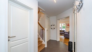 A hallway with a white door, a staircase, a wall-mounted light fixture, and a couch in the background. - Gode Stuv - Holiday home for up to 6 people with garden and sauna. (Sylt)