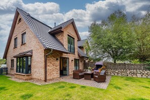 A two-story brick house with a sloped roof, a stone wall, and a patio with outdoor furniture. - Gode Stuv - Holiday home for up to 6 people with garden and sauna. (Sylt)