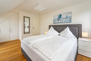 Un lit bien fait, avec du linge de lit blanc, un tableau représentant des voiliers accroché au mur et un luminaire moderne au plafond.