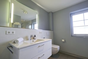 A modern bathroom with a white vanity, a large mirror, a toilet, and a window.