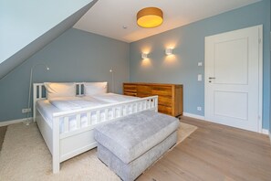 Una camera da letto con soffitto spiovente, un letto con biancheria bianca, una testiera in legno, due luci fissate al muro e una porta bianca.