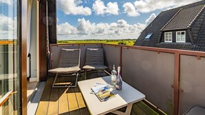 Un balcón con dos sillas acolchadas, una mesita con una revista y botellas, y vistas a un paisaje verde y una casa con techo inclinado.