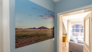 Un pasillo con un cuadro de paisaje grande en la pared, una puerta que da a una habitación con vistas al exterior y una pequeña zona de estar con una silla amarilla.