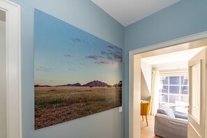 Un pasillo con un cuadro de paisaje grande en la pared, una puerta que da a una habitación con vistas al exterior y una pequeña zona de estar con una silla amarilla.