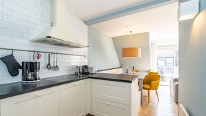 Una cocina moderna con armarios blancos, encimera negra y estufa empotrada. Hay una silla amarilla en la zona de comedor y un ventanal que proporciona luz natural.