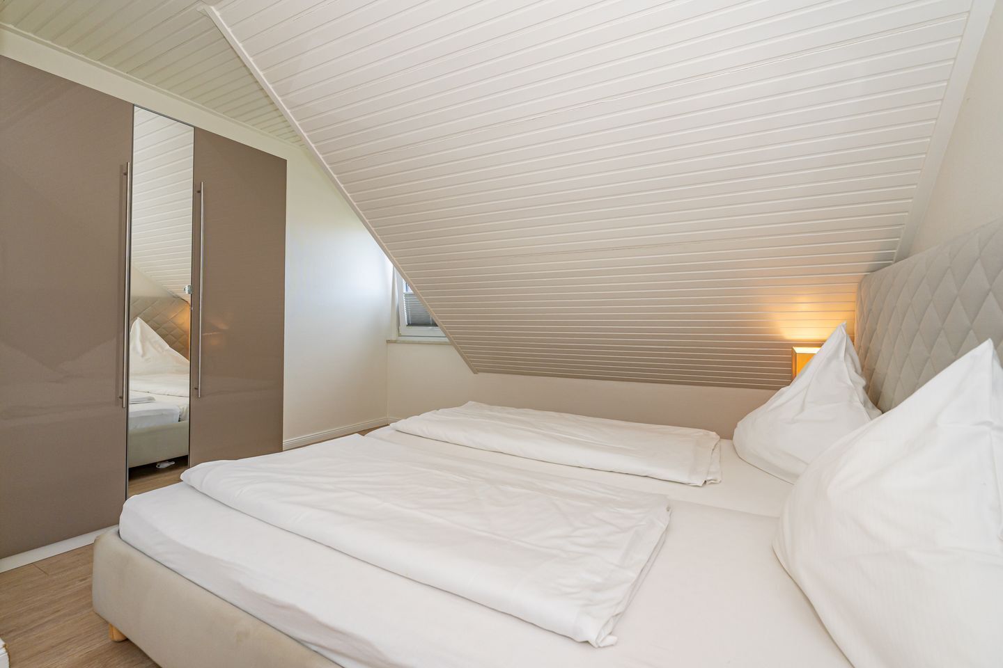 A bedroom with a sloped ceiling, a bed with white linens, a bedside lamp, and a wardrobe.