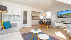A modern living room with a sofa, coffee table, and TV. There is a dining area with chairs and a table. The room has a kitchen in the background. - App.syltperle - Helle Wohnung Ruhig Gelegen in Westerland (Sylt)