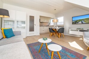 A modern living room with a sofa, coffee table, and TV. There is a dining area with chairs and a table. The room has a kitchen in the background. - App.syltperle - Helle Wohnung Ruhig Gelegen in Westerland (Sylt)