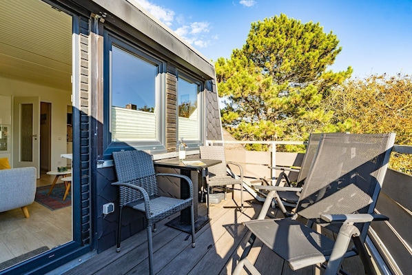 An outdoor patio with a table and chairs, a sliding glass door leading to a room, and a large tree in the background. - App.syltperle - Helle Wohnung Ruhig Gelegen in Westerland (Sylt)