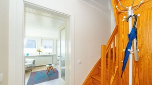 Un pasillo que conduce a una habitación con una escalera de madera, un paraguas azul colgado en la pared y vistas a una ventana con persianas.