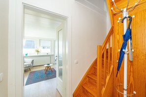 Un passadís condueix a una habitació amb una escala de fusta, un paraigua blau penjat a la paret, i una vista d'una finestra amb persianes.
