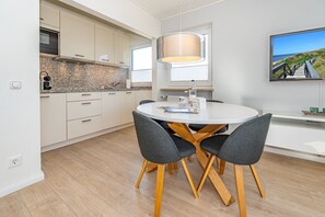 A modern kitchen with white cabinets, a granite countertop, and a built-in oven. There is a dining area with a round table and four chairs. - App.syltperle - Helle Wohnung Ruhig Gelegen in Westerland (Sylt)