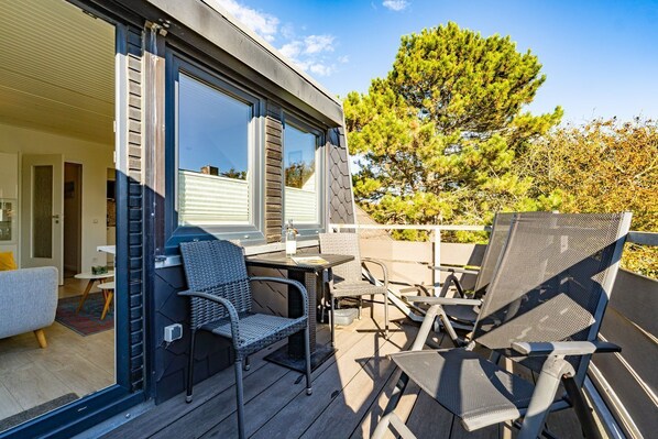 An outdoor patio with a table and chairs, a sliding glass door leading to a room, and a large tree in the background. - App.syltperle - Helle Wohnung Ruhig Gelegen in Westerland (Sylt)