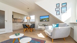 A modern living room with a grey sofa, a dining area with a table and chairs, a kitchen in the background, and a large window providing natural light. - App.syltperle - Helle Wohnung Ruhig Gelegen in Westerland (Sylt)