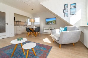 A modern living room with a grey sofa, a dining area with a table and chairs, a kitchen in the background, and a large window providing natural light. - App.syltperle - Helle Wohnung Ruhig Gelegen in Westerland (Sylt)