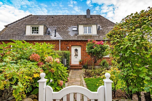 A two-story brick house with a white front door, a small front porch, and a garden with various plants and flowers.