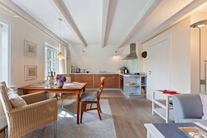 A modern kitchen with wooden cabinets, a dining area with wicker furniture, and a living room with a grey sofa. - App. Anne - Zentral Gelegene 2-zimmerwohnung, mit Gartennutzung (Sylt)