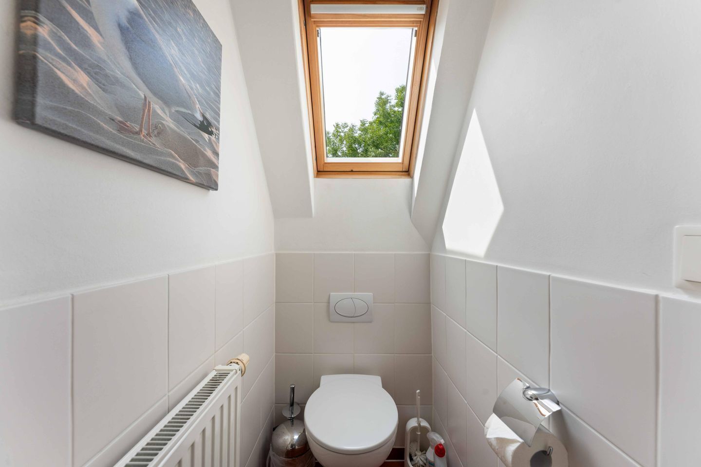 A compact bathroom with a skylight, white tiles, a toilet, a radiator, and a wall-mounted light fixture.