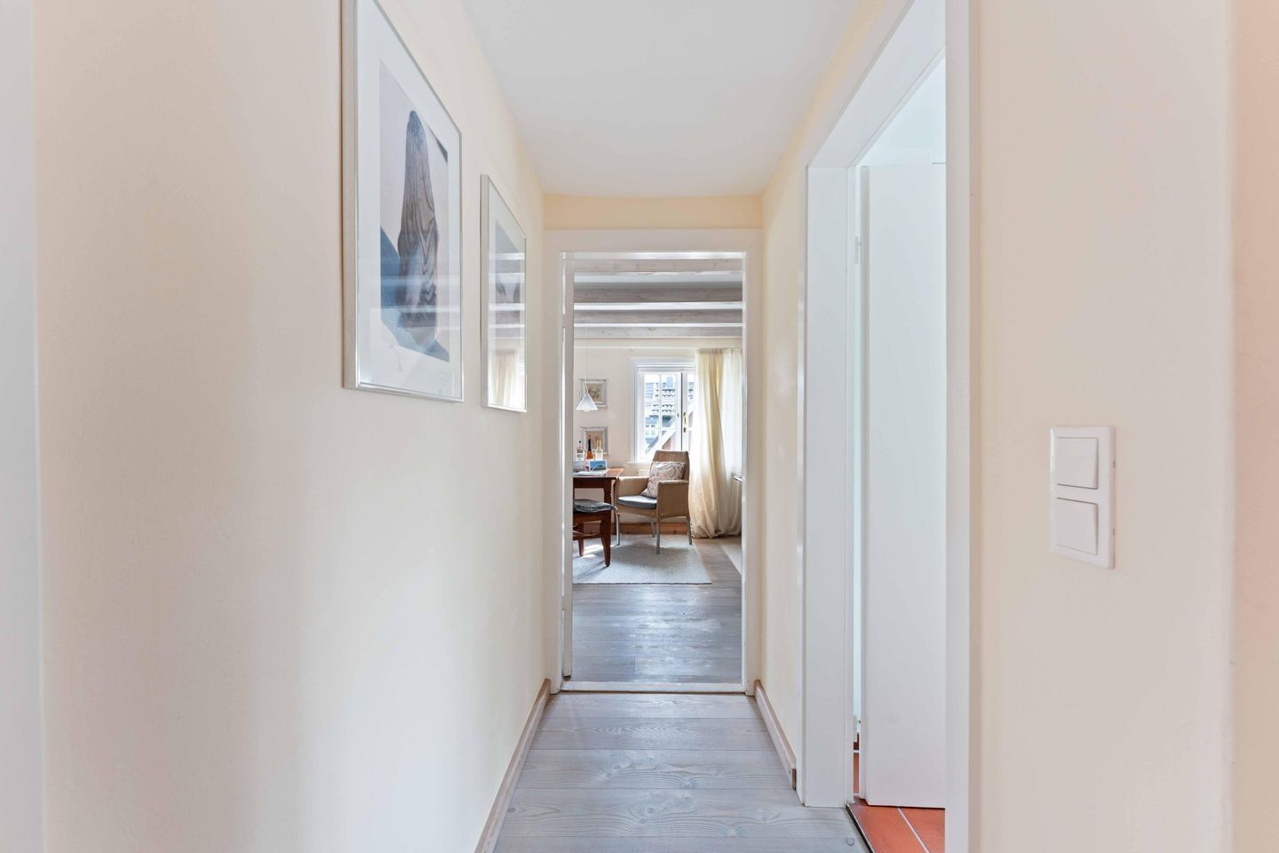 A hallway with framed artwork on the walls, leading to a room with a desk and chair.